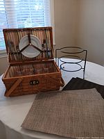 Woven picnic basket open on table showing striped fabric interior and metal plate holder next to 16 vinyl placemats.