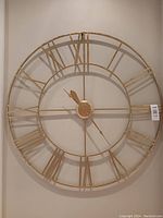 Front view of large gold-tone wall clock with Roman numerals mounted on a cream wall, showing entire clock face and hands.