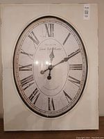 Front view of the large oval rustic metal clock without top bow and crown finial, showing its Roman numeral face and black hands.