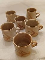Six handmade hand-thrown stoneware mugs with brown and gray abstract brushstroke design, varying slightly in size and shape, arranged on white surface.