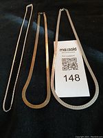 Three flat chain necklaces displayed on dark fabric background with auction label showing lot number 148. From left to right: gold-tone herringbone chain, silver-tone herringbone chain, silver-tone Boston link chain.