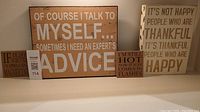 Four word art items displayed on a shelf: two rectangular plaques and two square fridge magnets with quotes about happiness, advice, aging, and humor.