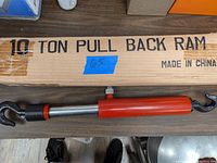 Photo showing red hydraulic pull back ram resting on a table with its original cardboard box behind it. The box is labeled '10 TON PULL BACK RAM' and 'MADE IN CHINA'.
