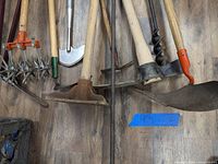 Overview of assorted lawn and yard tools spread on wooden floor including rakes, hoes, shovel, auger, and cultivators.