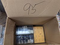 Top-down view of a cardboard box containing two plastic compartment boxes and a yellow labeled box filled with assorted washers, rubbers, and fasteners.