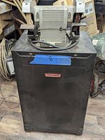 Front view of the black metal cabinet with the mounted bench grinder on top, showing KREFTORNIK label on cabinet and power cord.