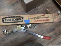 Craftsman winch puller tool and original box placed on wooden floor, showing metal build, hooks, red handle, and branding on box.
