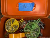 View of open orange utility box showing multiple round plastic containers with lead fishing weights and loose lead weights inside