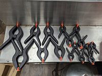 Photo shows 11 black plastic spring clamps with orange tips of varying sizes arranged on a metal surface. Sizes include extra large, large, medium, and small.