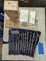 Photo of Sears Craftsman folding chisel and plane blade sharpener original box, set of drill bits in fabric case and two plastic cases containing small drill bits and metal parts on table.