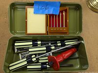 Green metal utility box opened showing multiple screwdrivers with cream and black handles alongside a wooden box set of precision screwdrivers.