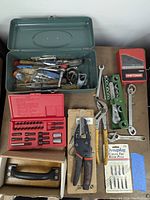 Layout of all items on table including drill bit set in red Craftsman case, various hand tools, wire stripper, tire repair kit, and rubbing brick.