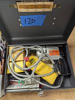 Photo of an electric drill inside a gray metal carrying/storage case, with power cord and yellow accessory holder visible.