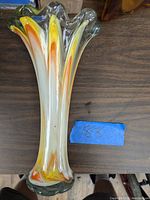 Side view of tall glass vase showing multicolored vertical streaks (orange, yellow, green) and fluted splayed top design on wooden surface.