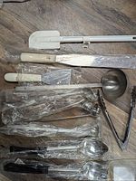 Photo of assorted white handled spatulas, large knife, metal ladle, tongs, and other wrapped utensils on floor.