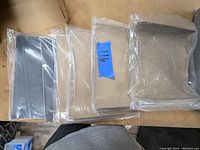 Multiple sealed packages of sandpaper sheets sorted by grit, some labeled with blue tape showing grit numbers like 111 and 116