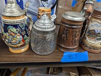 Photo showing six beer steins lined up on shelf displaying traditional and thematic designs, various materials and colors