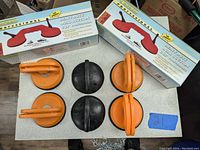 Top-down photo showing six suction cups with handles, three black and three orange, placed on a table with their boxes behind them.