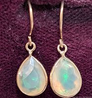 Front view of pair of 14K gold earrings with pear-shaped faceted opal gemstones resting on burgundy fabric.