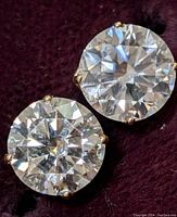 Close-up of two round-cut moissanite stones in 14K yellow gold four-prong settings on a dark velvet background.