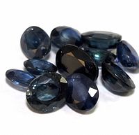 Group of 11 oval-shaped faceted sapphire gemstones in various shades of deep blue, arranged closely on a white surface.