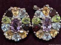 Close-up image of a pair of sterling silver earrings with a central violet gemstone surrounded by pear-shaped yellow, light blue, green, and deep red gemstones.
