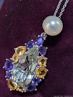 Close-up view of the pendant showing the large faceted pear-shaped green amethyst surrounded by oval and round amethyst and citrine stones with a white pearl above, resting on a dark purple velvet background.