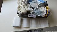 Photo showing the box of drapery hooks in plastic packaging, multiple metal hooks visible, white ribbing strips rolled and partially laid out