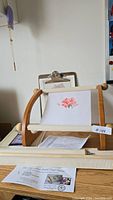 Wide shot of the wooden lap stitch frame holding a fabric with rose embroidery, showing the setup on a table with pattern sheets.