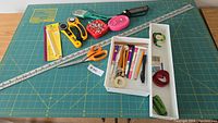 Full view of sewing supplies on cutting mat including rulers, rotary cutters, scissors, pins, and box of markers.