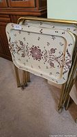 View of the three vintage metal TV trays stacked with one tray dented and with maroon floral pattern on cream background and the four gold-tone metal folding stands, one with wheels.