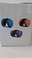 Box showing images of three color options: blue, orange, and purple dog ear muffs with black adjustable straps.
