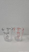 Two heatproof shot glasses placed side by side, one with tablespoon and teaspoon measurements in black print, the other with ounce and milliliter measurements in red print, both with handle.