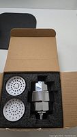 Open box showing chrome shower head filter unit and two white circular spray plates with multiple holes, set in black foam padding.