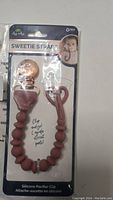 Photo of new silicone pacifier clip in dusty rose color with beaded strap and metal clip attached, on retail packaging card labeled 'Sweetie Strap'.