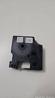 Single vinyl label tape cartridge showing front and label details, black plastic housing with white label.