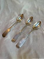 Three antique forged silver spoons laid out on a white fabric, showing the front side with monogram engravings on the flat handles.