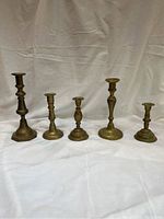 Five brass candlesticks in a row showing front and side views with a plain backdrop