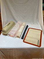Tan ceramic bread loaf pan, two translucent white Tupperware nonstick pastry rolling mats with red markings, dark oven liner roll, beige Silpat baking sheet with brown edge.