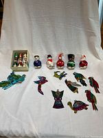 Wide view showing three wooden wisemen in a box, five colorful glass snowmen figurines in costumes, eight enameled tin ornaments depicting birds, angels, and fish, and two firebird decorations on a white fabric background.