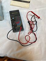 Micronta digital multimeter with attached red and black test probes and instruction manual in background.