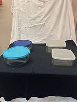Four glass storage containers with plastic lids on black and white background, showing front view of containers and lids, different shapes and lid colors
