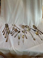 Wide view of all mixed silver plated serving utensils laid out on white cloth background.