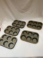 Five metal muffin tins arranged side by side on a white cloth background showing the tops with six muffin cups each.