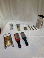 Front view of the lot showing three can openers, a bottle lid opener, and four graters of various sizes and designs arranged on a white fabric background.