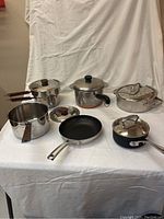Cookware pieces including double boilers, saucepans with wood handles, and frying pans, displayed on white cloth background.
