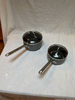 Four aluminum nonstick sauce pans with glass and metal lids arranged on white fabric backdrop. Shows wear and condition of pans and lids.