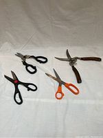 Four kitchen shears with black, orange, and wooden handles arranged on white fabric.