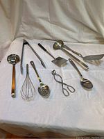 Nine stainless steel kitchen tools including ladles, tongs, spoons, spatulas and a whisk arranged on a white cloth.