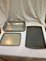 Three metal baking sheet pans displayed on white cloth backdrop: a large heavy metal pan, a smaller Cuisinart pan, and a large rimmed pan.